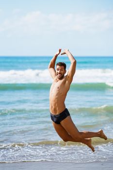 Funny young man with beautiful body in swimwear jumping on a tropical beach.