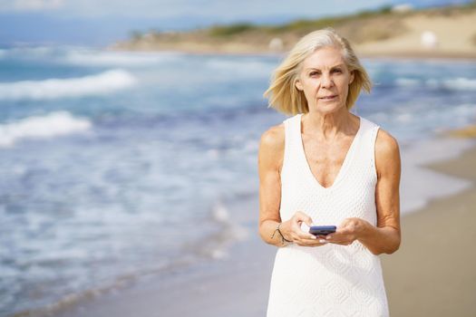 Mature woman walking on the beach using a smartphone. Elderly female enjoying her retirement at a seaside location.