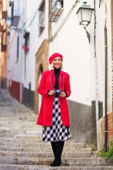 Full body of confident young female, tourist in fashionable outfit with photo camera, standing on old stairs in town and smiling while exploring city historic district in daytime