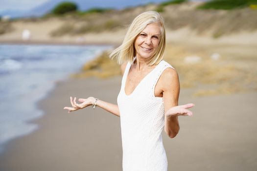 Smiling mature woman opening her arms on a tropical beach. Elderly female enjoying her retirement walking on the beach.