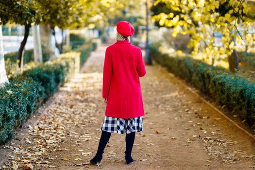 Full body back view of anonymous female in red outfit standing on pathway in park with trees on autumn day