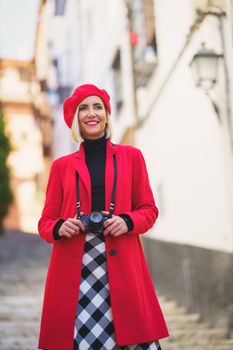 Low angle of happy young lady with blond hair, in stylish warm clothes and beret, smiling and looking away while admiring and photographing town on camera