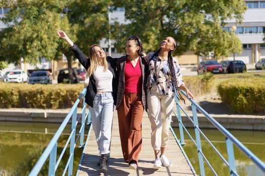 Full body of cheerful multiracial female friends strolling together on wooden footbridge over river in city on sunny summer day
