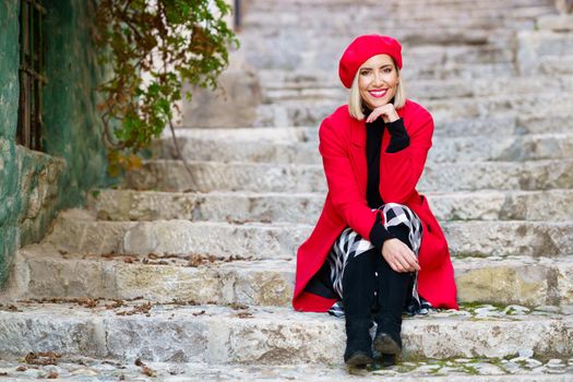 Full body of confident young lady, with blond hair and red lips in elegant coat and beret, sitting on shabby stone stairs with hand at chin and smiling in town