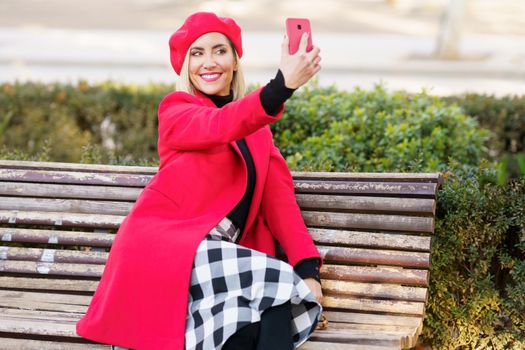 Delighted female in stylish red outfit taking self portrait on smartphone while sitting on wooden bench in park on autumn day