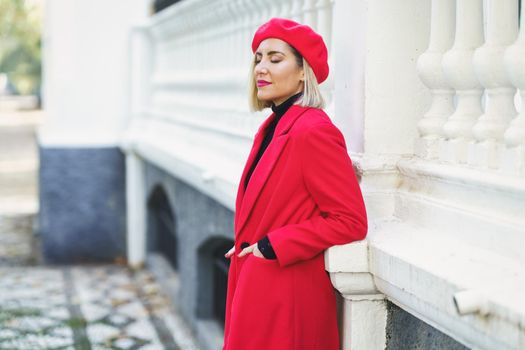 Side view of attractive female in red outfit with beret standing with hands in pockets near white fence on street