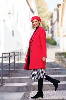 Full body of trendy female in red beret looking at camera while strolling on stone staircase on street of city