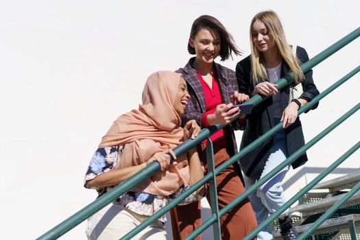 Low angle of joyful young Muslim woman in hijab, laughing while watching photo on smartphone with happy female friends, standing together on metal staircase on sunny day