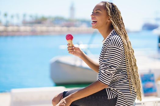 Beautiful young black woman eating a strawberry ice cream while enjoying the view of the seaport.