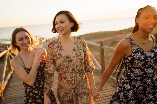 Optimistic multiracial female friends holding hands and walking together on wooden boardwalk while spending time together on beach against sea