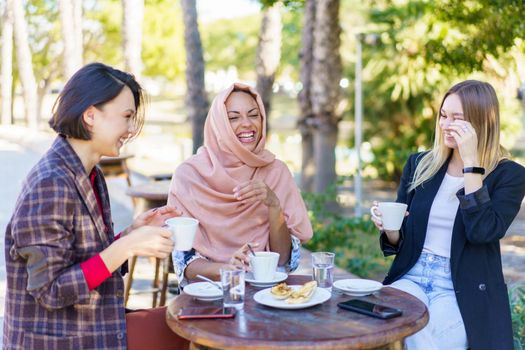 Happy multiethnic female friends laughing while sitting at table with snack and coffee on terrace of cafeteria on summer day