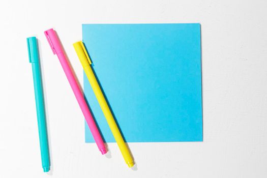 Multi-colored paper blue square on a white background, three multi-colored ballpoint pens. Office concept. Place for text