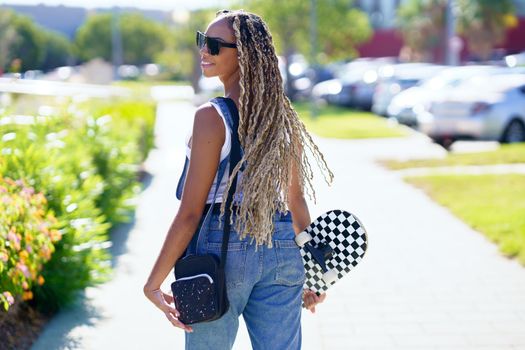 Back view of positive black female skater with braids and skateboard walking along street in summer and looking over shoulder