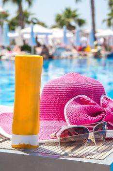 Bottle of sunscreen, hat and sunglasses next to swimming pool in hotel with palms on background