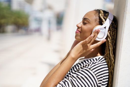 Side view of glad African American female, with closed eyes, touching headphones and listening to music, while leaning on wall on sunny day on blurred background of city street