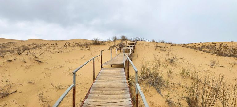 The staircase rises to a sandy dune. Climbing to the top in the desert. Sarykum dune. Dagestan, Russia. A unique sandy mountain in the Caucasus on a cloudy day. Grass grows on a sand dune.