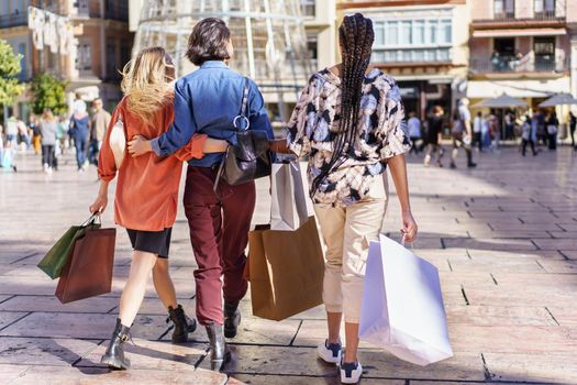 Back view of unrecognizable trendy young female friends in stylish outfits cuddling while walking on city square with paper bags after successful shopping