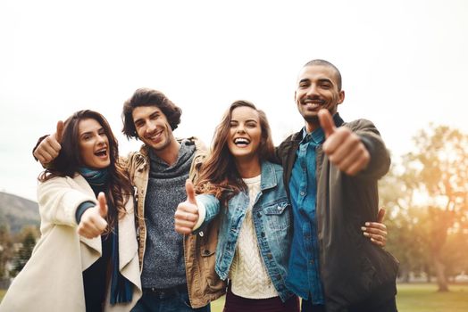 Portrait of a group of cheerful young friends huddled together while showing thumbs up outside during the day.