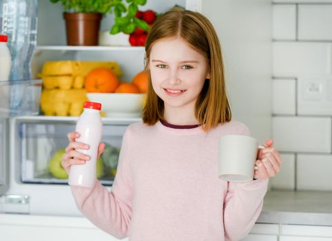 Pretty preteen girl holding yogurt and cup mug at kitchen at home and looking at camera. Beautiful female kid with milk products smiling