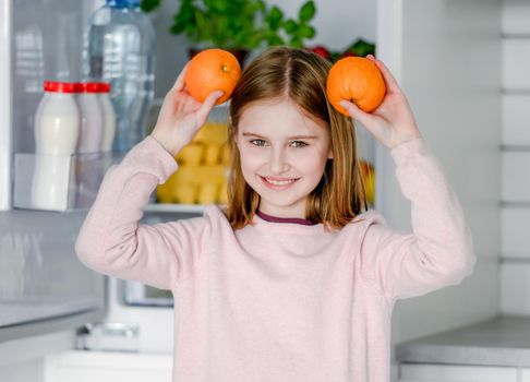 Pretty preteen girl holding oranges near head and playing with citruses at kitchen at home. Beautiful female kid with fruits