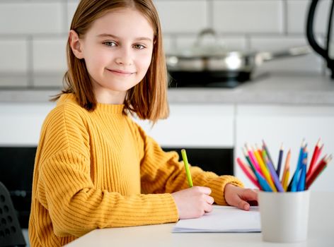 Preteen girl painting with colorful pencils at home, looking at camera and smiling. Pretty kid child drawing sitting at table portrait