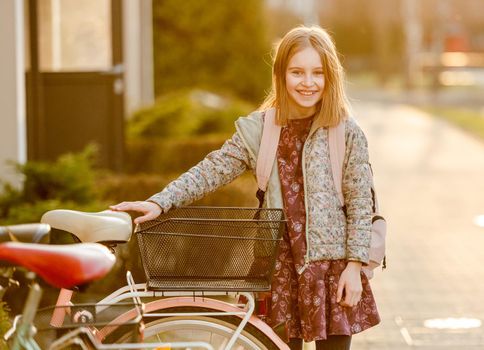Preteen girl child with bycicle looking at camera and smiling in beautiful sunset light. Pretty kid walking outdoors with vehicle