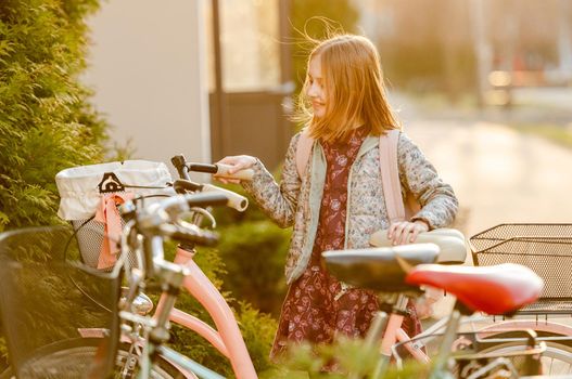 Preteen girl child looking at bycicle and smiling in beautiful sunset light. Pretty kid walking outdoors with bike