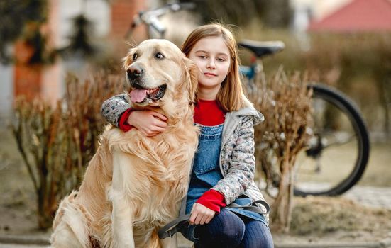 Preteen girl with golden retriever dog sitting in park in beautiful spring autumn day. Adorable female child kid hugging purebred doggy pet outdoors portrait