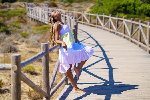 African female wearing a beautiful dress on a boardwalk on the beach. Fashion woman outdoors.