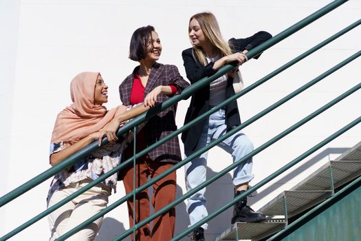 Low angle of trendy young multiethnic women best friends, on stylish outfit and hijab, leaning on metal railing while standing on staircase and looking away in city on sunny day