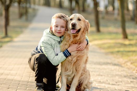 Preteen boy with golden retriever dog sitting outdoors and smiling at springtime in sunny day. Child kid with doggy pet in park
