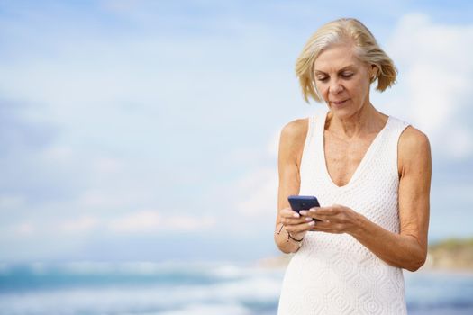 Mature woman walking on the beach using a smartphone. Elderly female enjoying her retirement at a seaside location.