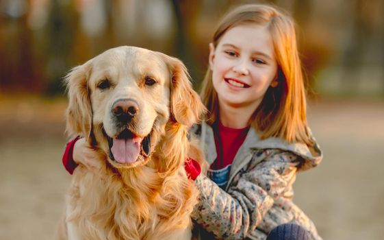 Preteen girl with golden retriever dog sitting in park in beautiful spring autumn day. Adorable female child kid hugging doggy pet outdoors and smiling portrait