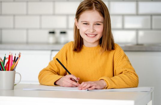 Preteen girl painting with colorful pencils at home, looking at camera and smiling. Pretty kid child drawing sitting at table portrait