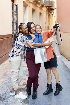 Full length of glad multiracial female friends taking selfie on camera while standing on street near building after shopping