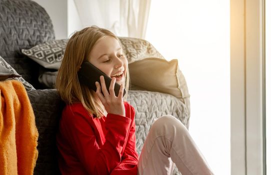 Preteen girl holds smartphone in her hand and smiles. Cheerful child spends time sitting on floor talking by mobile phone