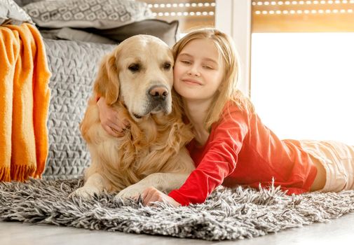Cheerful child girl with golden retriever dog lies on floor near sofa and smiling hugging pet. Happy preteen girl with purebred labrador doggy