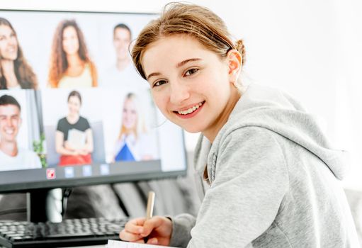 Girl during remote educaton class via video link with friends classmates on display. Teenager e-learning on distance with computer at home in coronavirus pandemic time