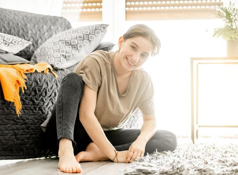 Teenager girl sits on floor and smiles at home. Happy young female person in cozy interior with daily light