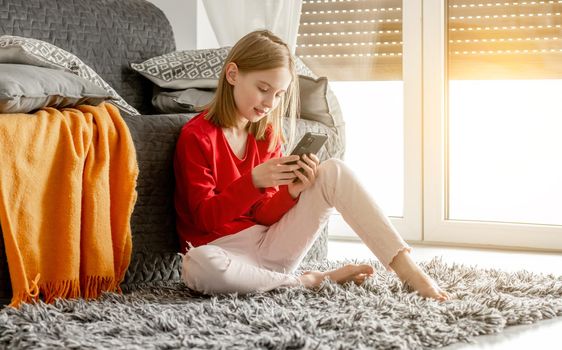 Child girl holds smartphone in her hand and smiles chatting with friends. Cheerful kid spends time with cellphone sitting on floor near sofa by window.