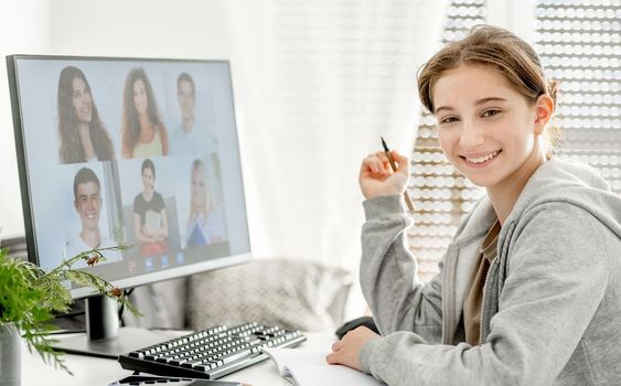Girl during remote educaton class via video link with friends classmates on display. Teenager e-learning on distance with computer at home in coronavirus pandemic time