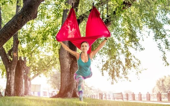 Sport girl practicing fly yoga in hammock at nature and stretching her body standing on one leg. Active woman during aero gymnastics outdoors in summer