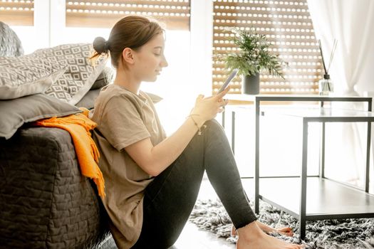 Pretty girl sits with smartphone near sofa at home. Female teenager person spends time with cellphone chatting on floor next to window.