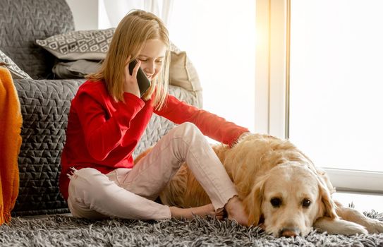 Cheerful child girl sits with golden retriever dog on floor and talks by smartphone. Happy preteen kid stroking purebred dog labrador during communication by cellphone