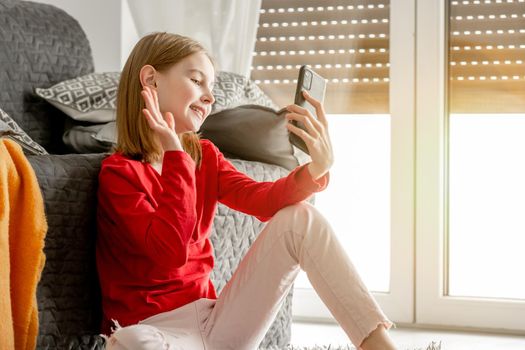 Smiling child girl communicates by smartphone and waves her hand. Cheerful kid spends time talking by video on celphone sitting on floor near window.