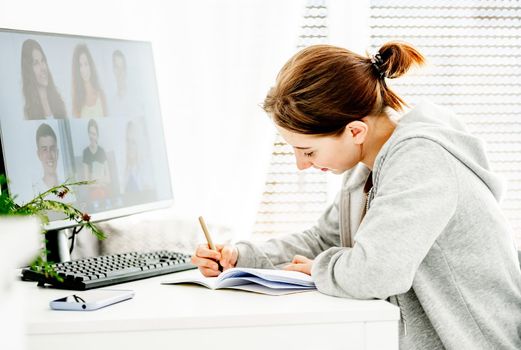 Girl writing in notebook during remote educaton class via video link with friends classmates. Teenager e-learning with computer at home in coronavirus pandemic time