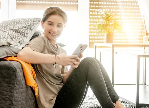 Pretty girl sits with smartphone near sofa at home. Female teenager person spends time with cellphone chatting on floor in cozy light room