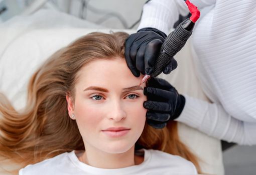 Beautiful blond girl with blue eyes during eyebrows microblading process. Permanent makeup closeup view from above