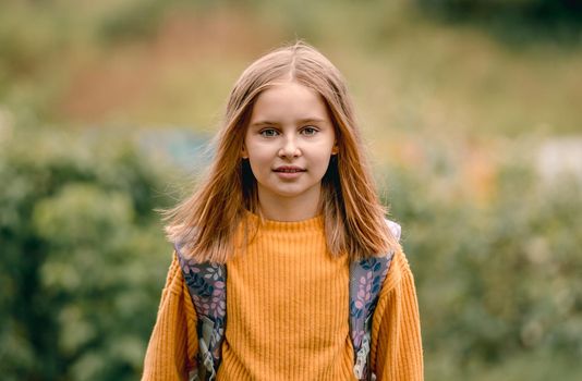 Cute school girl with packpack in the park portrait. Pretty female kid with beautiful eyes looking at camera and smiling outdoors