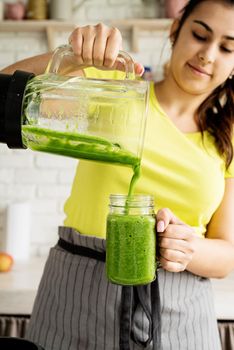 Healthy eating, dieting concept. Young brunette woman making green smoothie at home kitchen, poring from blender to jar